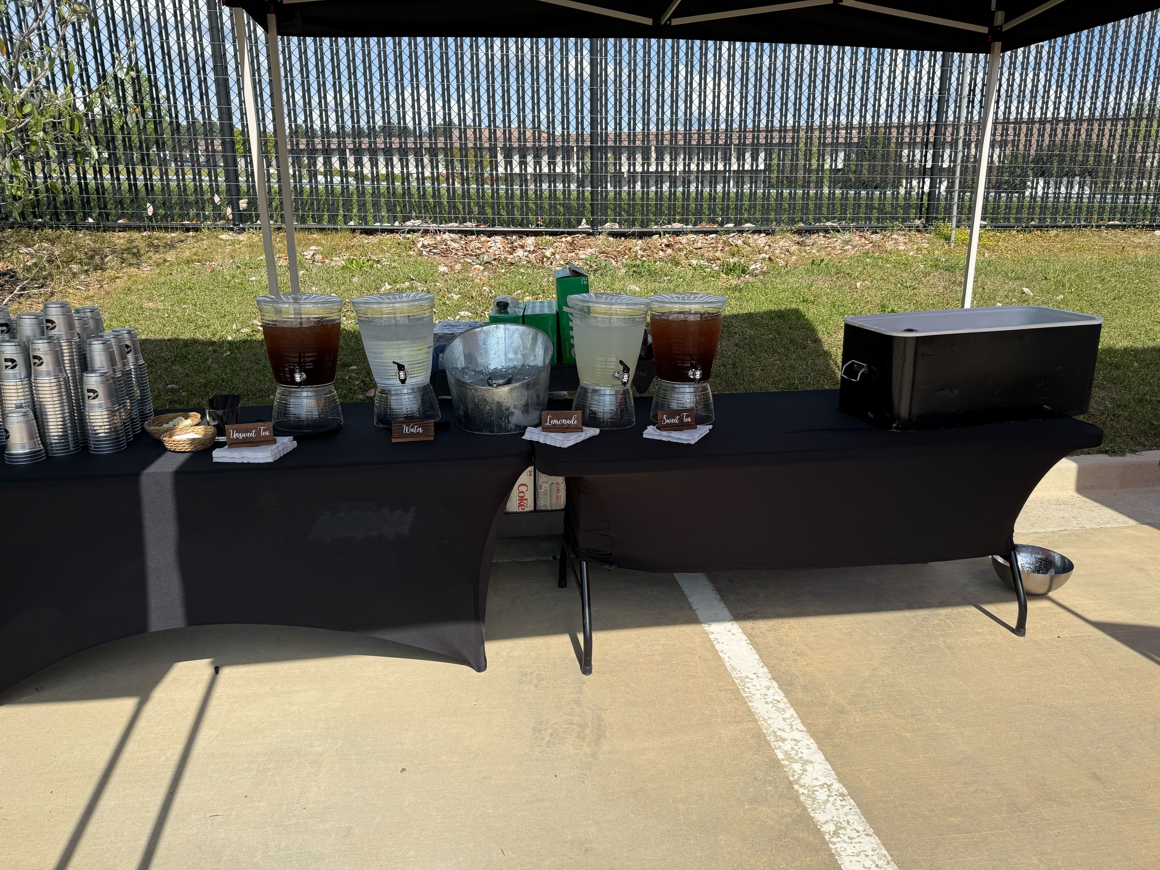 Outdoor beverage station with labeled dispensers for unsweetened tea, water, lemonade, and sweet tea on a black tablecloth — Atlanta corporate event catering