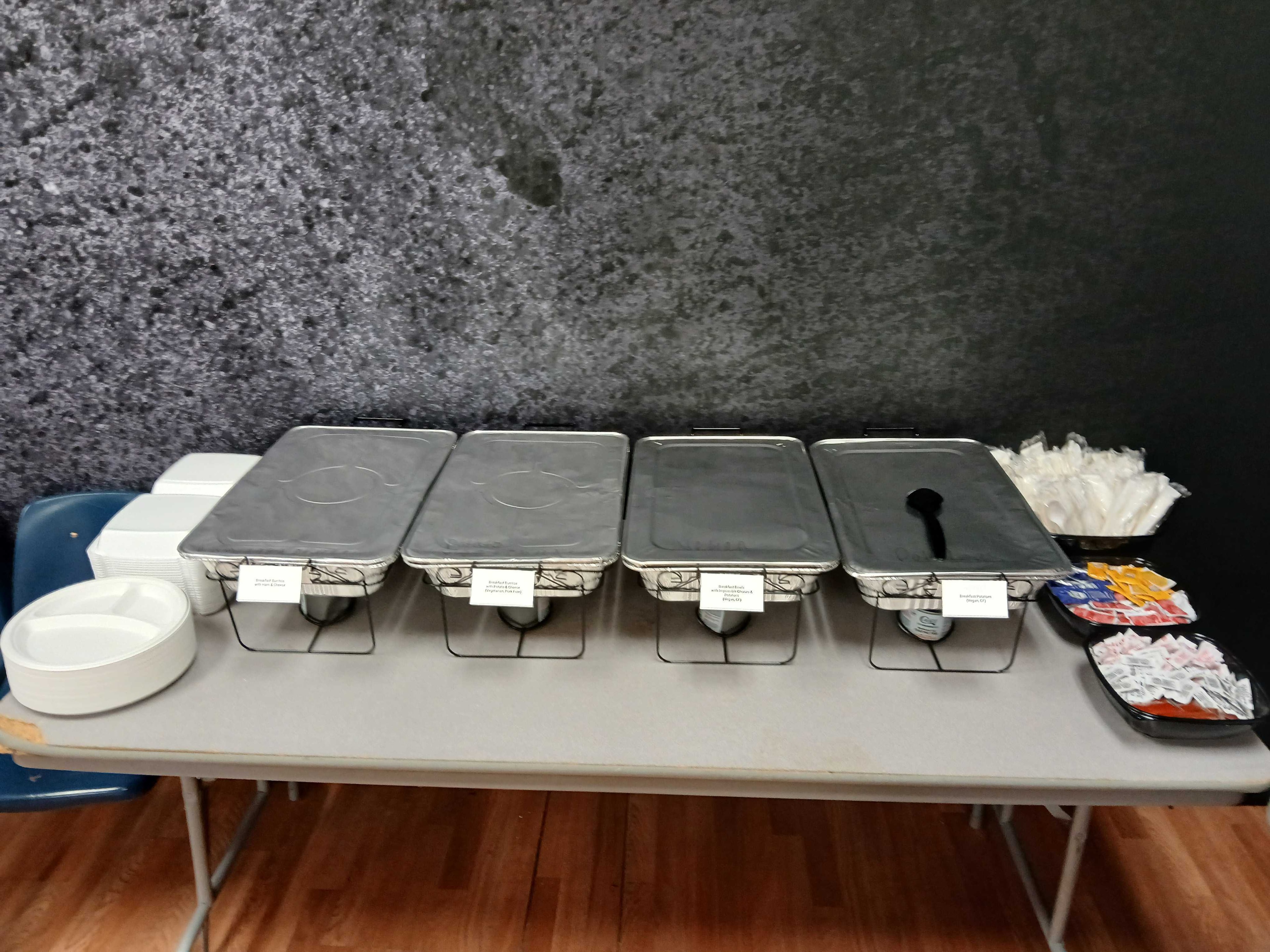 Four labeled chafing dishes with condiments and utensils on an Atlanta film production catering table, dark textured wall