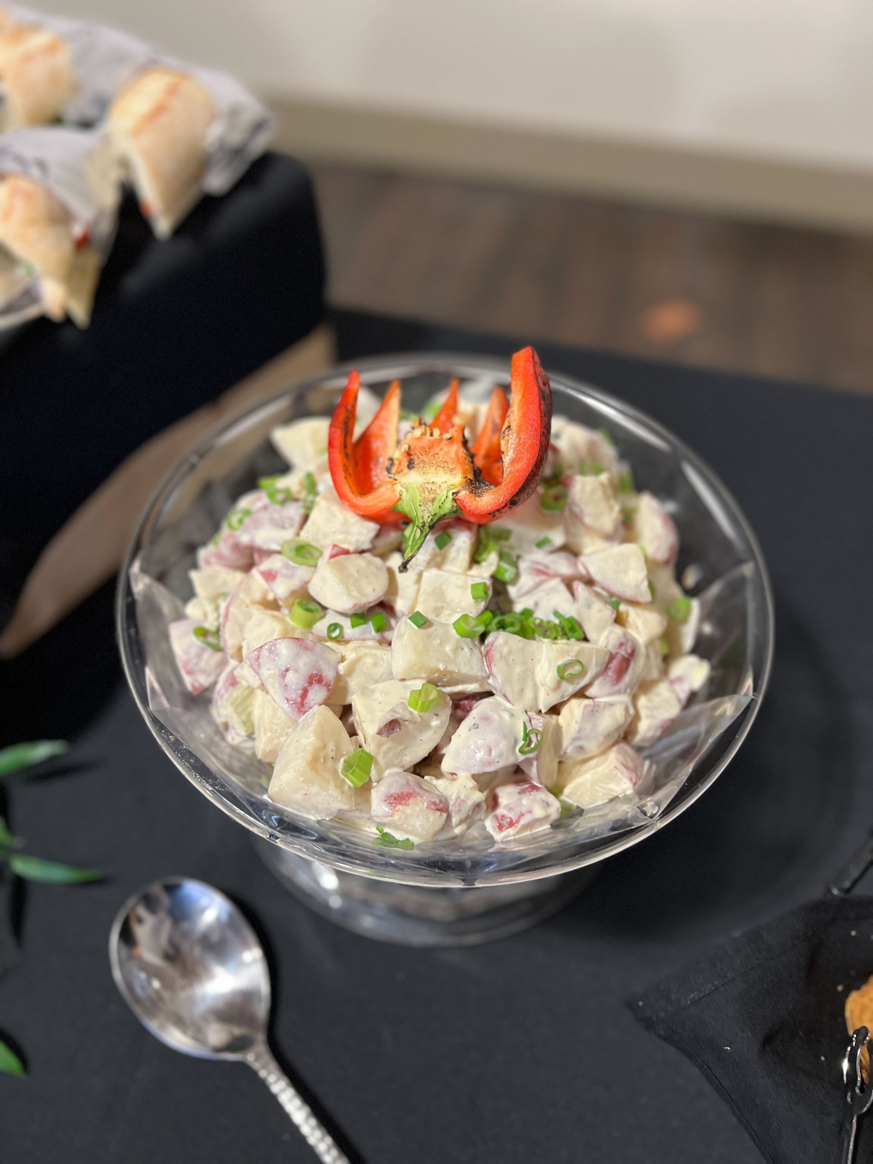 Creamy red potato salad in a glass pedestal bowl garnished with a roasted red pepper flower and sliced green onions — Atlanta production catering side dish