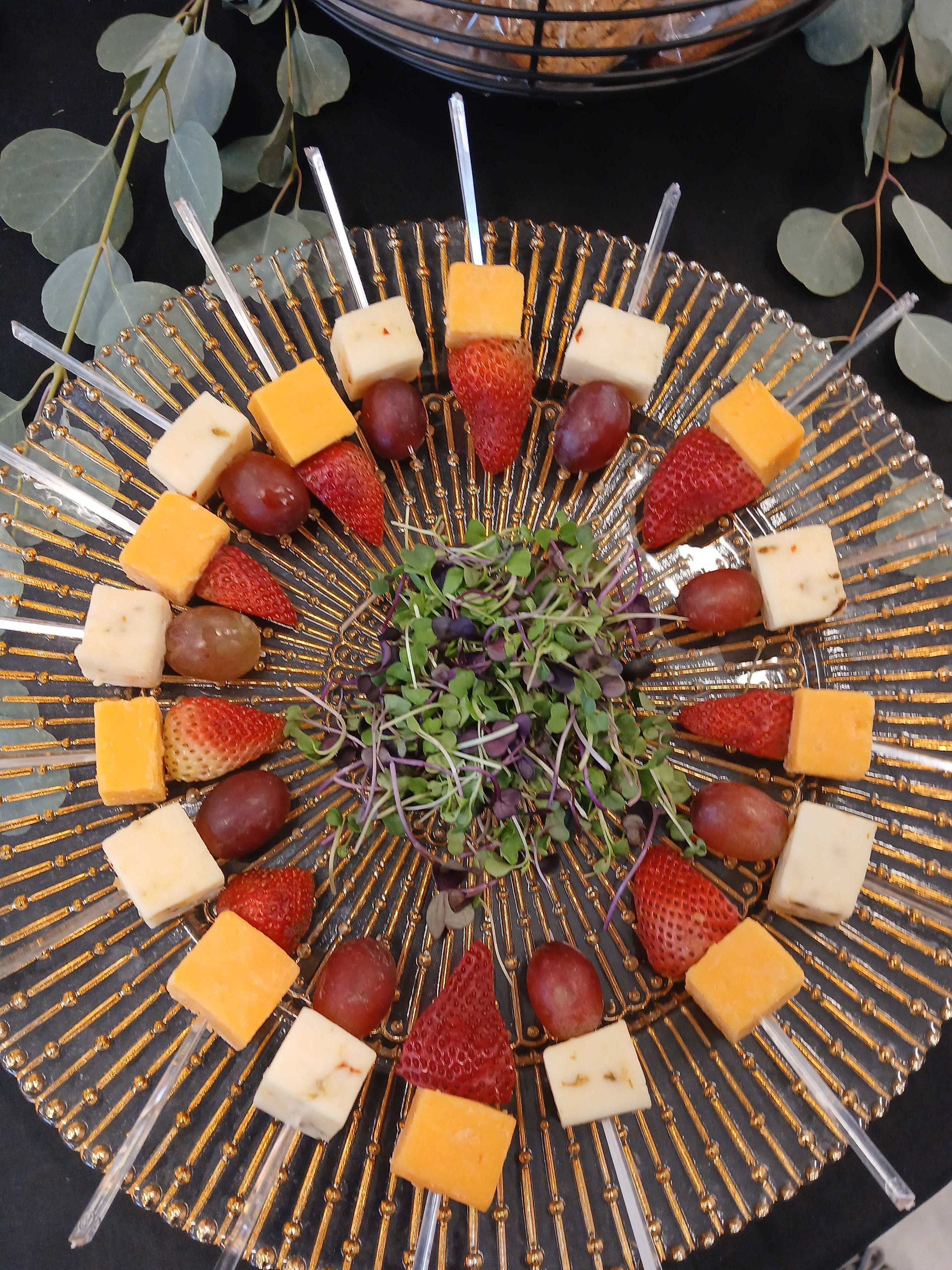 Cheese and fruit skewer platter with cheddar, pepper jack, strawberries, red grapes, and microgreens on a gold-rimmed glass plate with eucalyptus — Atlanta event catering