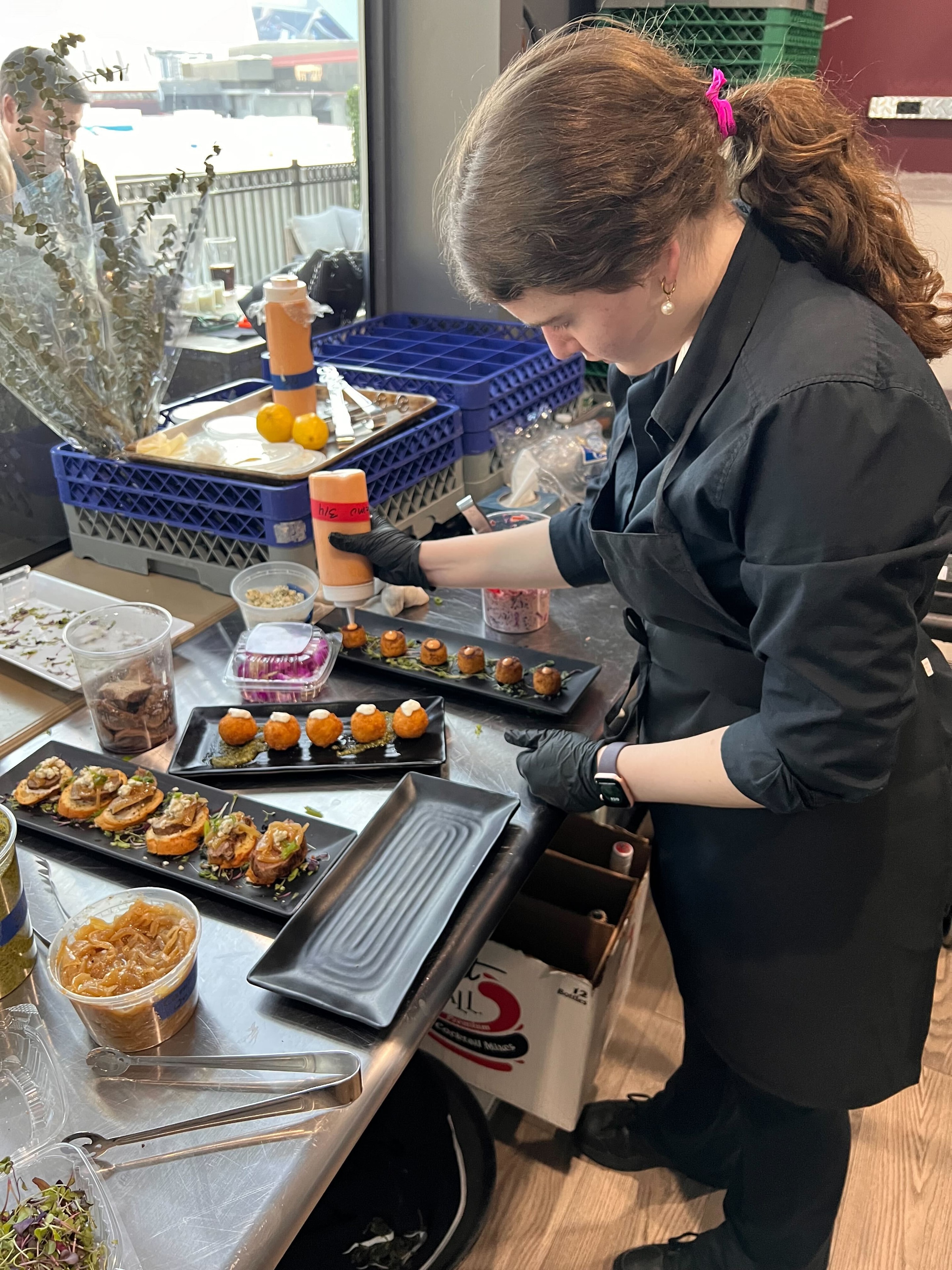 Atlanta Craft Services chef in black uniform plating golden arancini and bruschetta on black rectangular serving plates at a catering prep station