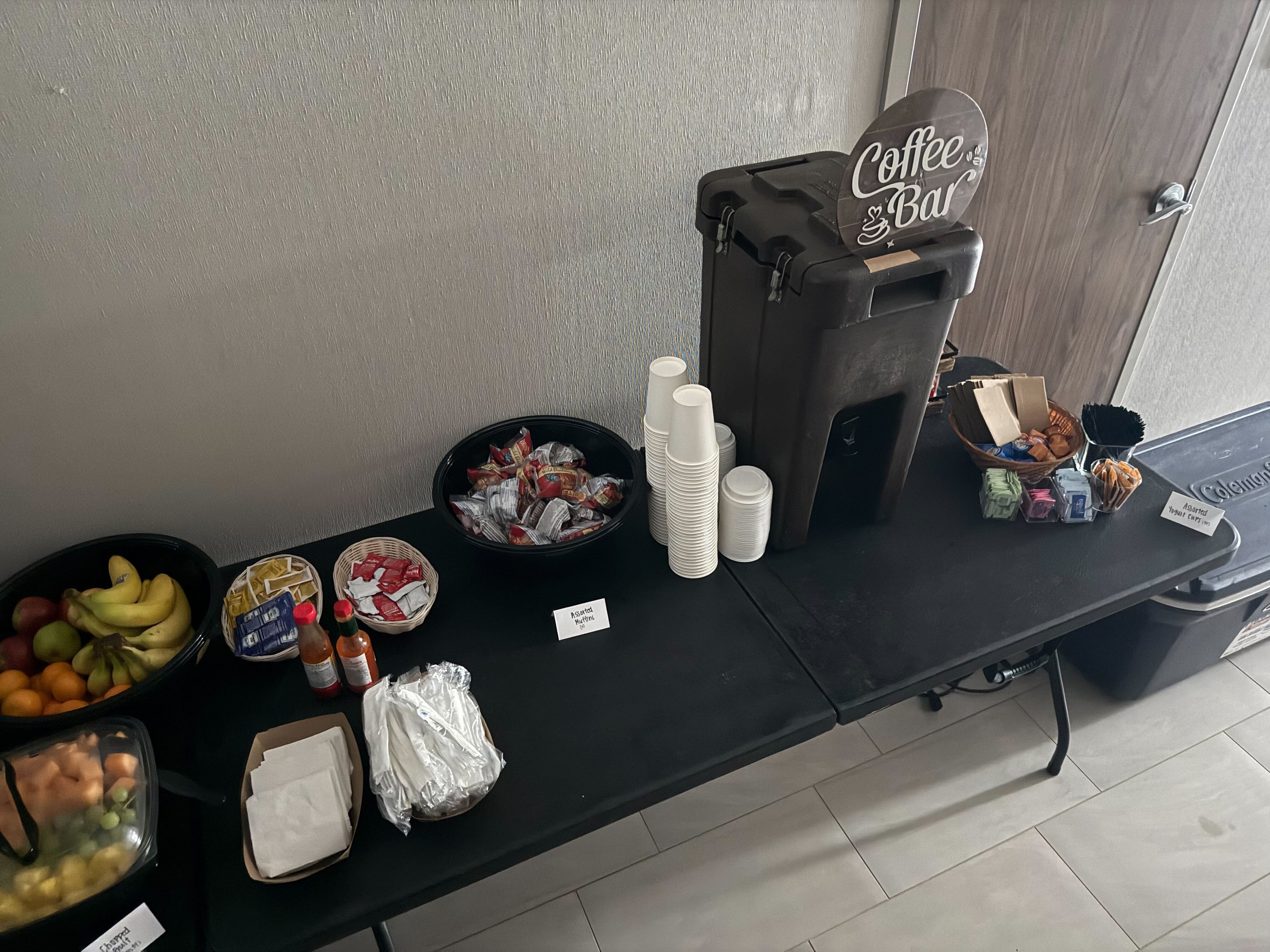 Atlanta Craft Services coffee bar with insulated dispenser, Coffee Bar sign, fresh fruit, snacks, muffins, and condiments on a black table