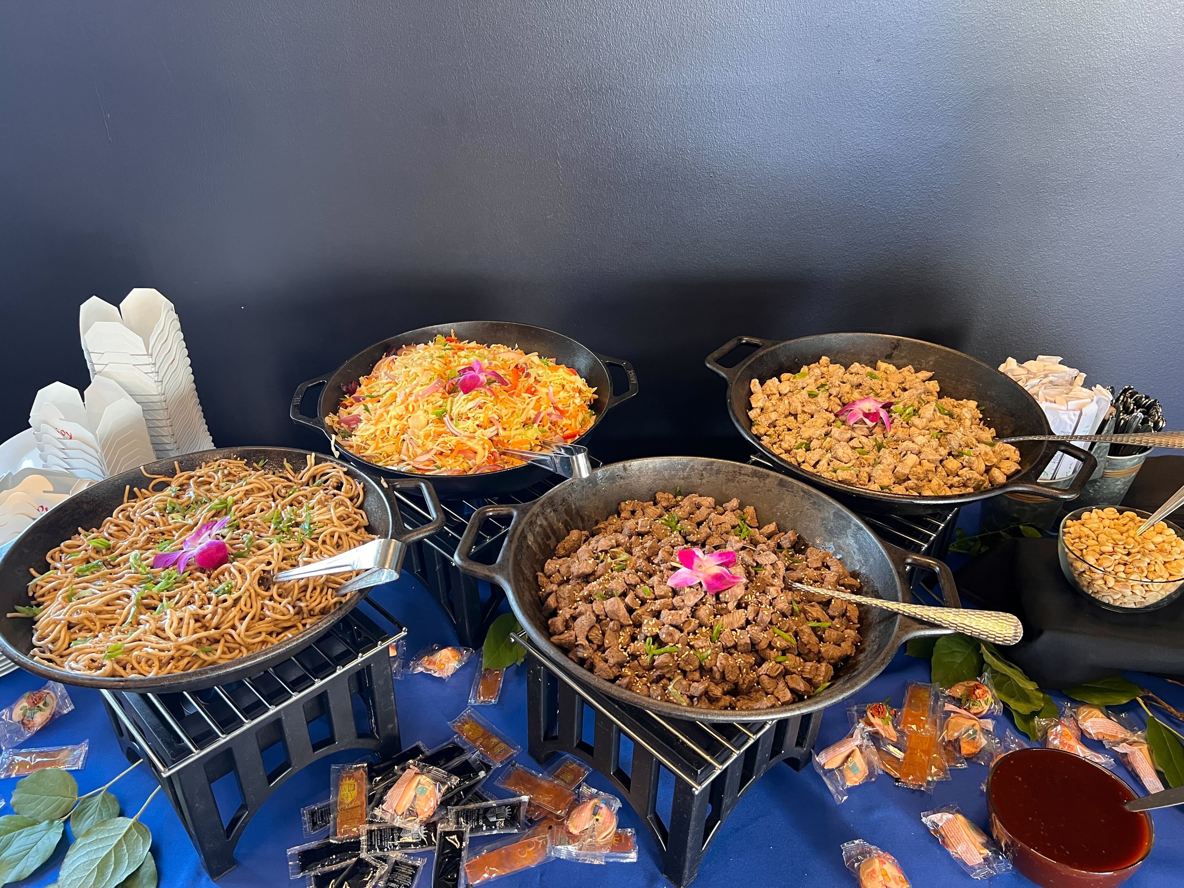 Atlanta corporate event Asian-inspired buffet with cast-iron woks of lo mein noodles, fried rice, sesame beef, and crispy chicken on black risers with orchid garnishes and takeout boxes