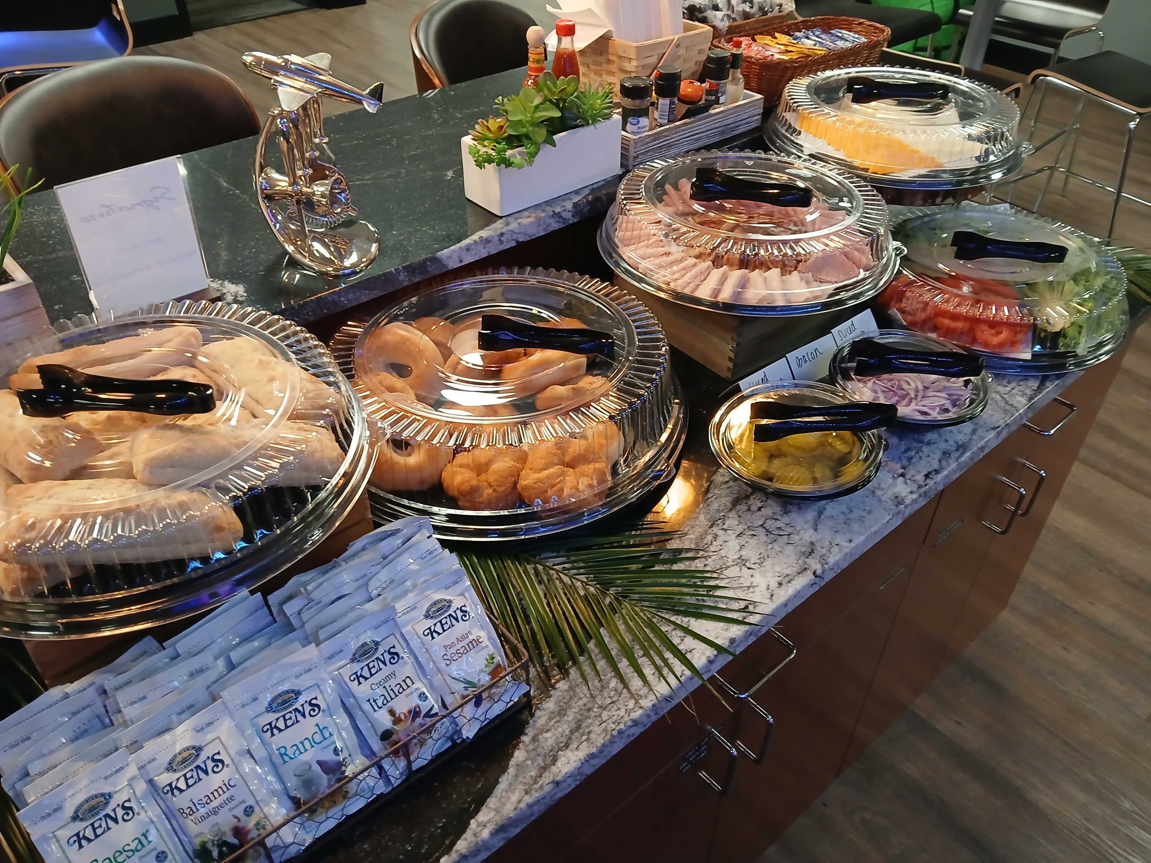 Corporate catering deli buffet on a dark granite counter with bagels, rolls, deli meats, tomatoes, pickles, and Ken's dressing packets — Atlanta