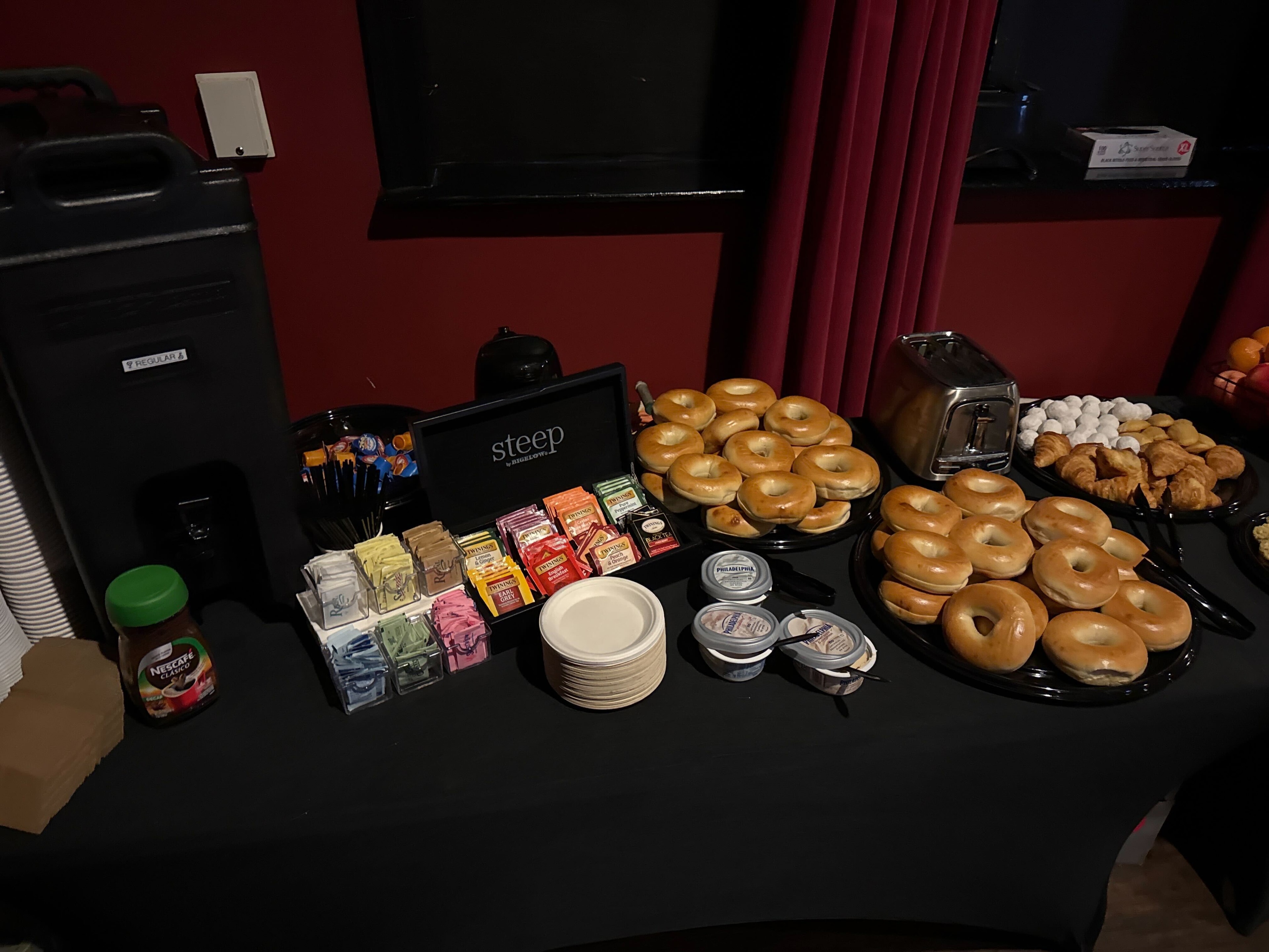 Craft service bagel and coffee station with Cambro dispenser, Steep tea display, assorted bagels, cream cheese, and croissants on a black tablecloth — Atlanta film set
