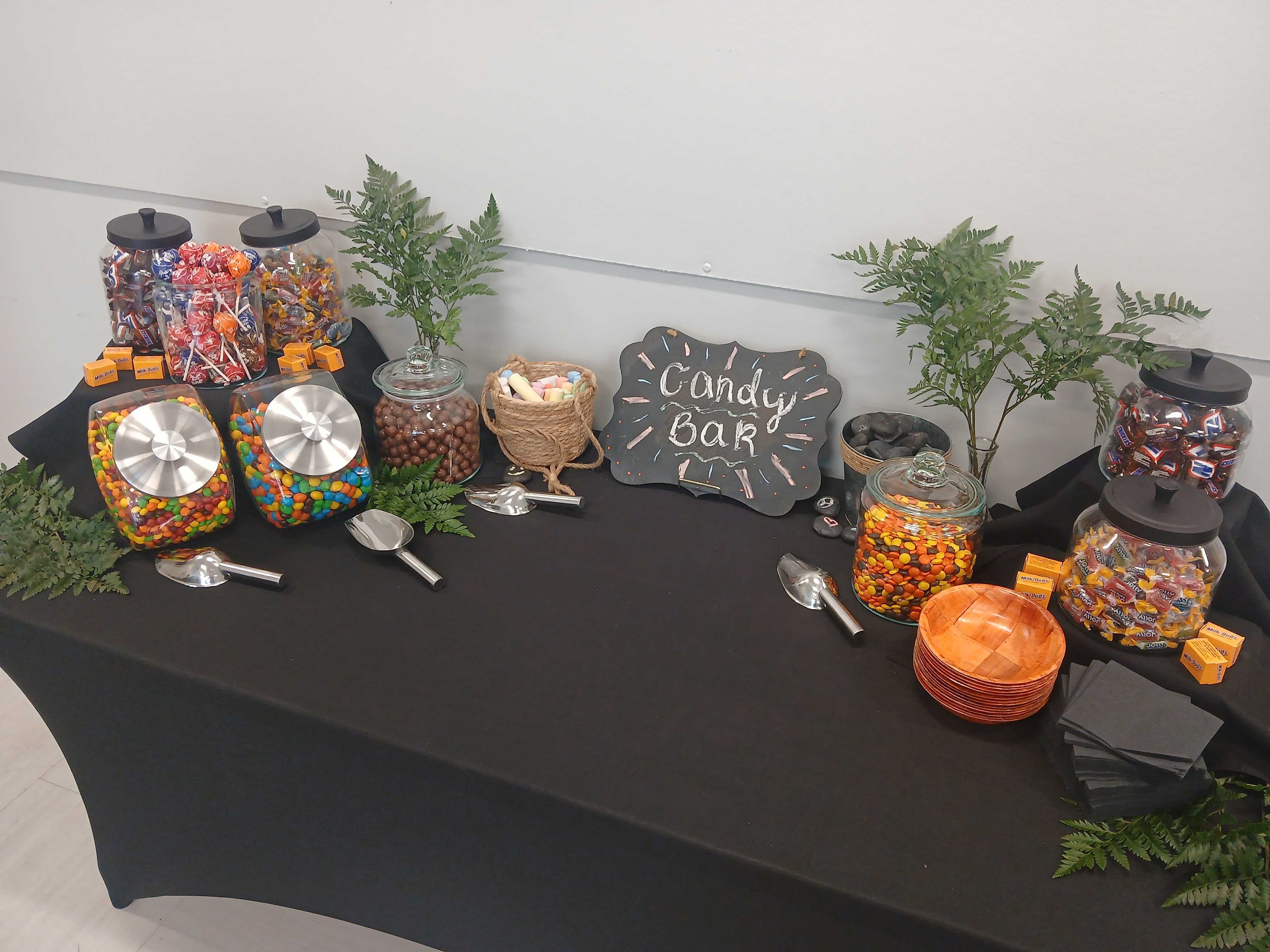 Atlanta craft service candy bar display with glass jars of M&Ms, Reese's Pieces, lollipops, chocolate, and Milk Duds on a black tablecloth with fern greenery