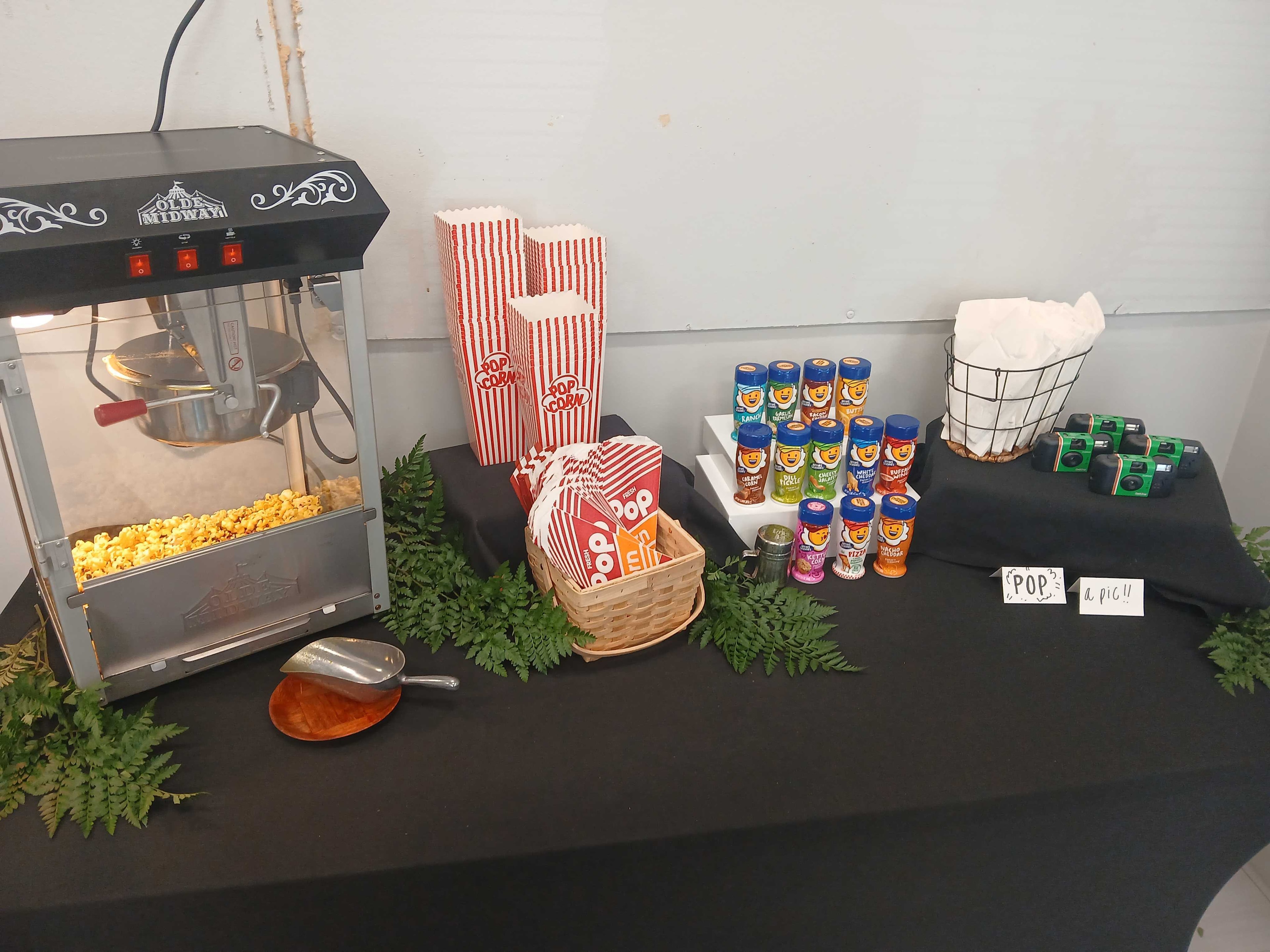 Atlanta craft service popcorn station with Olde Midway machine, striped popcorn bags, Kernel Season's seasonings, and fern greenery on a black tablecloth