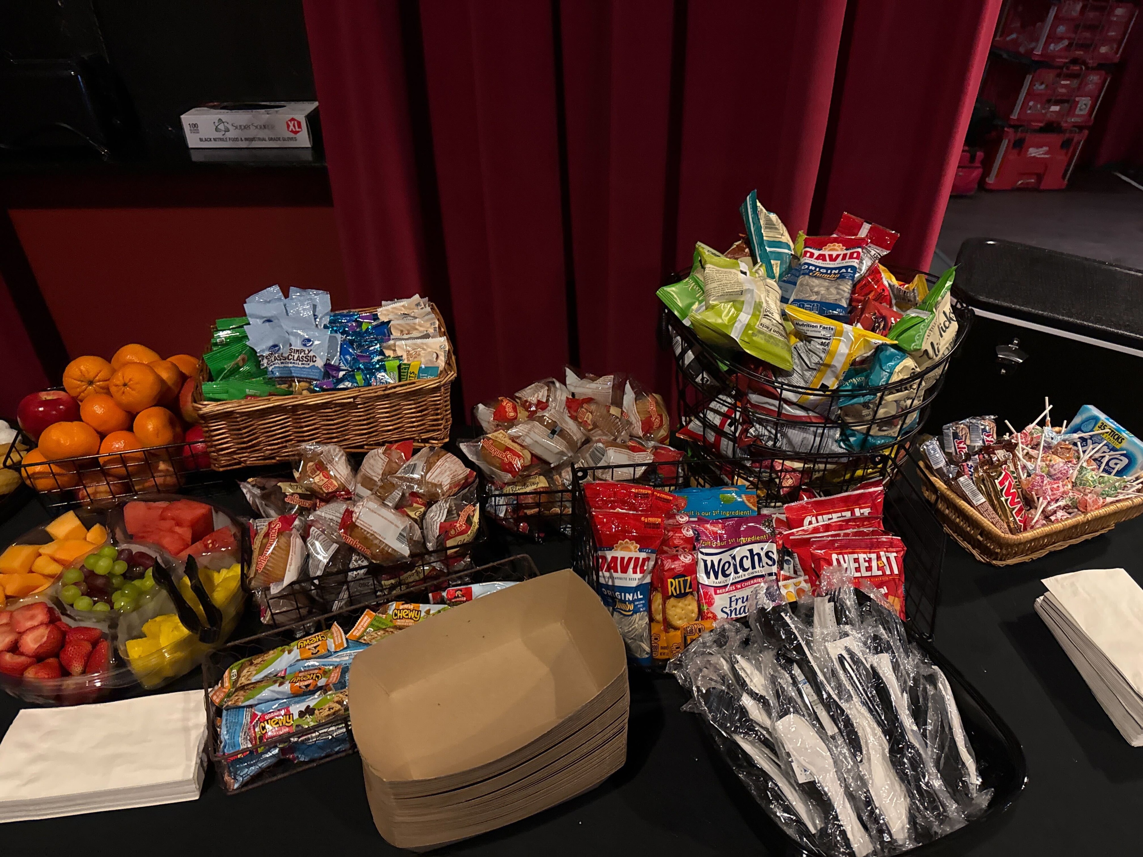 Atlanta craft service snack station with wire baskets of chips, packaged snacks, fresh fruit, and a fruit platter on a black tablecloth against a red wall