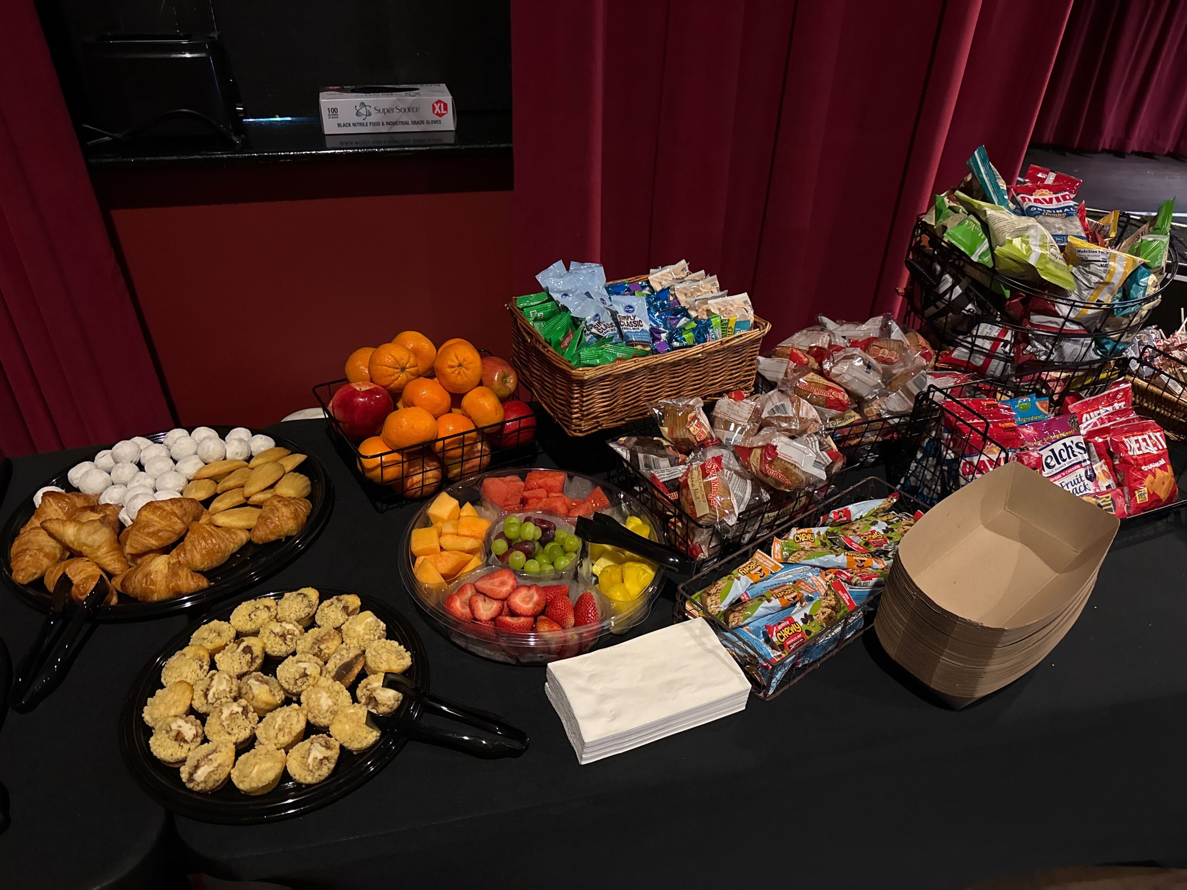 Atlanta craft service snack table with croissants, fruit platter, packaged snacks, wire baskets, and fresh fruit on a black tablecloth against a red wall
