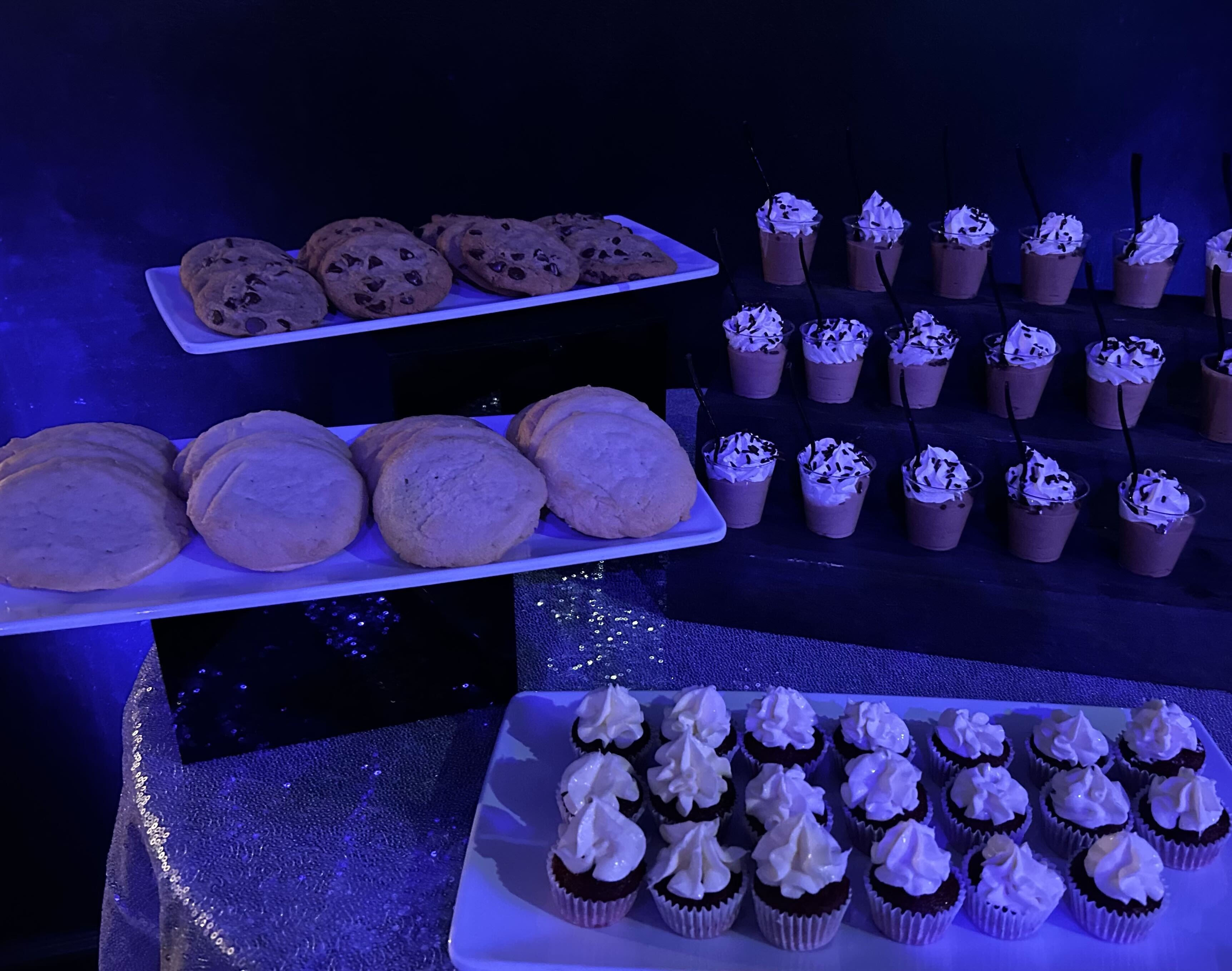 Atlanta corporate event dessert display — chocolate chip cookies, sugar cookies, chocolate mousse cups, and red velvet cupcakes under blue event lighting