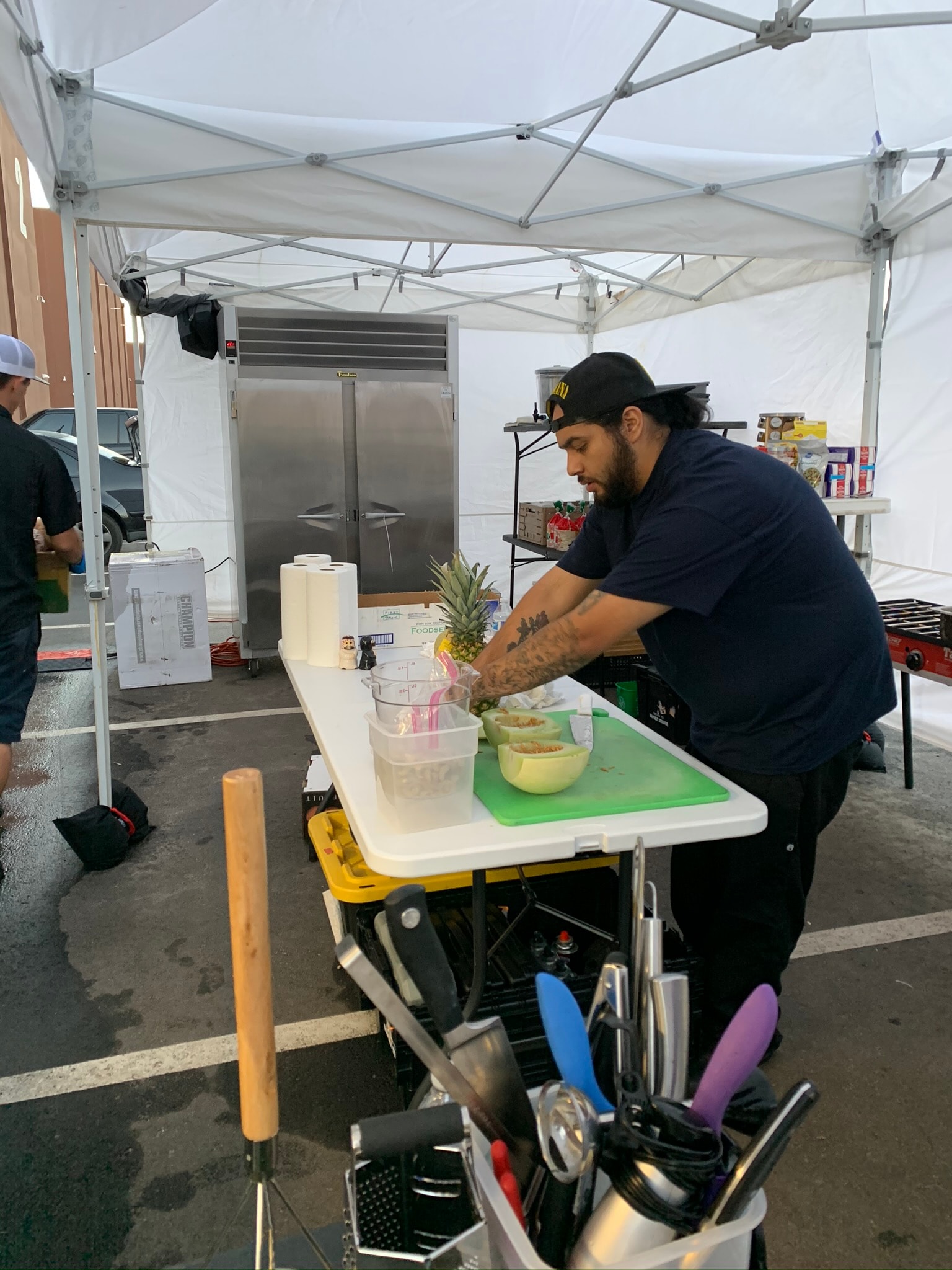 Chef cutting a pineapple at an outdoor prep station under a tent with a full knife roll and commercial refrigerator in the background — Atlanta production catering