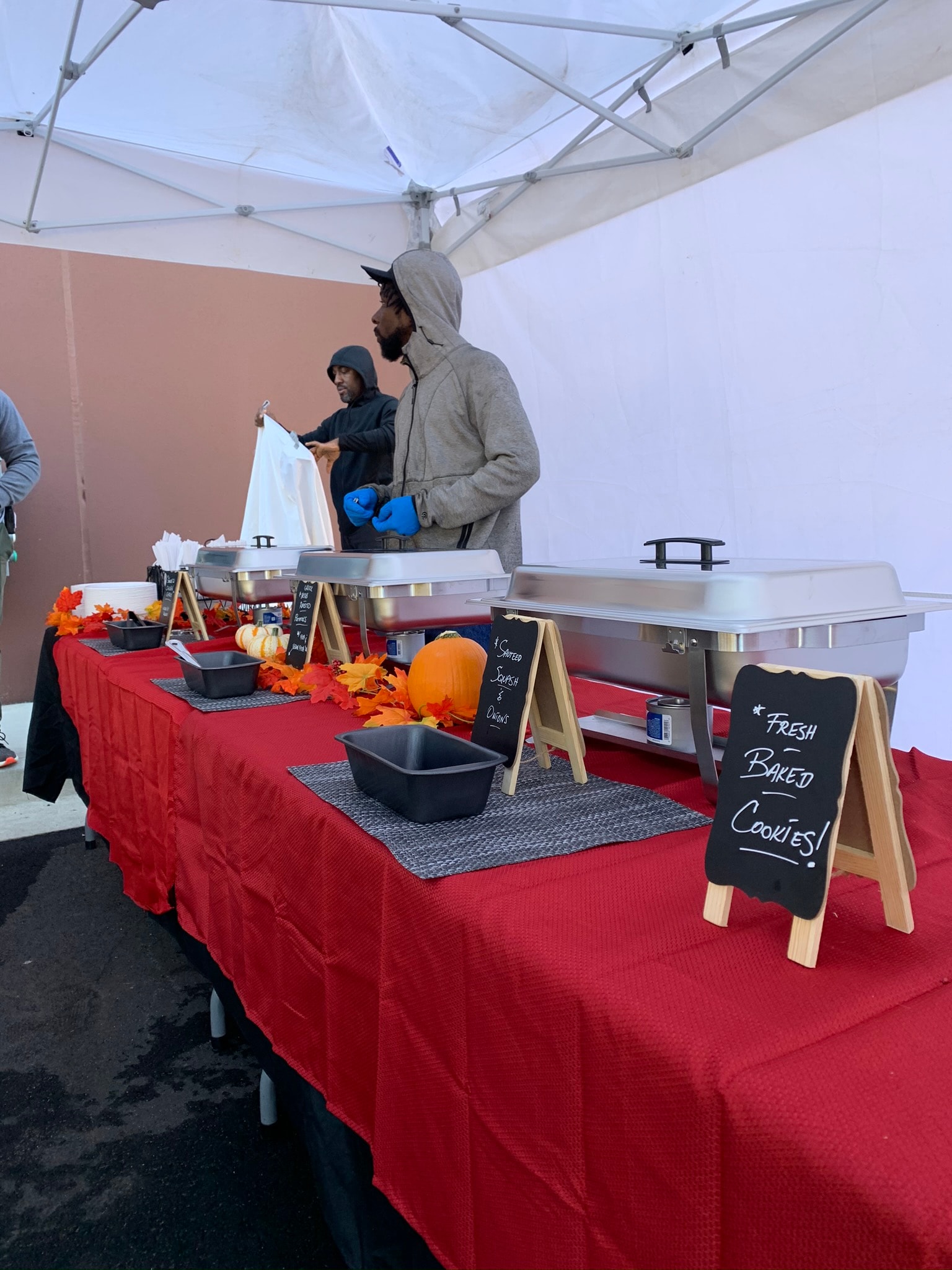 Atlanta Craft Services crew setting up a fall-themed buffet under a white tent with red tablecloth, chafing dishes, autumn leaf decorations, pumpkins, and a "Fresh Baked Cookies" chalkboard sign