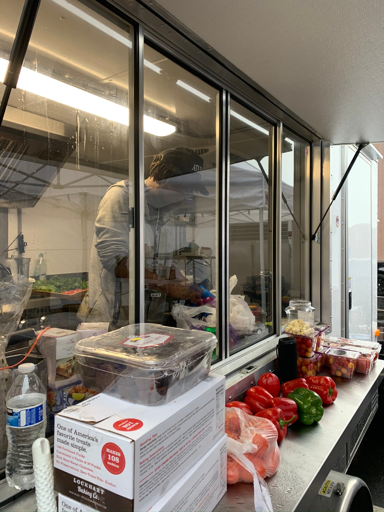 Daytime view of chef working inside a mobile kitchen trailer through glass windows, with fresh produce and Lockhart Baking Co. cookie dough boxes on the counter