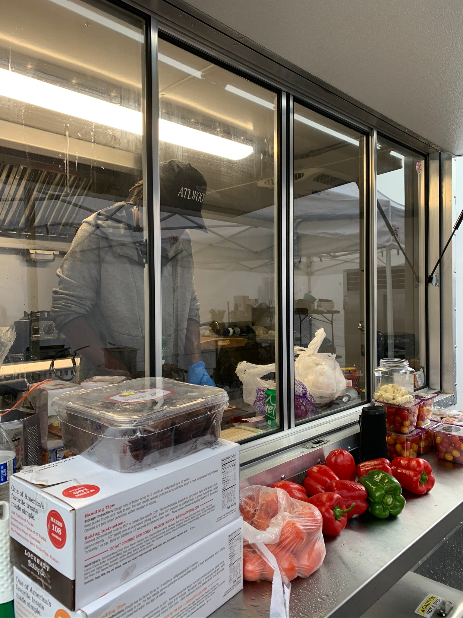 Chef working inside a mobile kitchen trailer viewed through glass windows, with fresh red and green bell peppers and cherry tomatoes on the counter outside