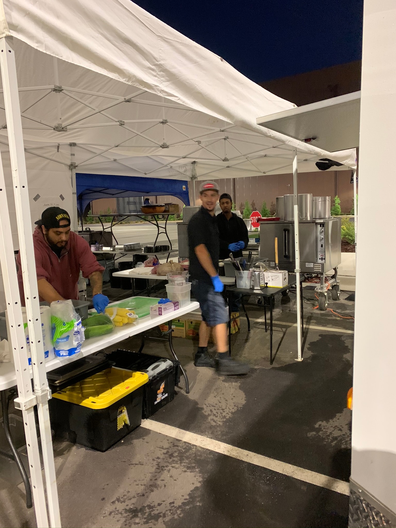 Three Atlanta Craft Services crew members prepping food under a white tent at night with commercial kitchen equipment, prep tables, and yellow toolboxes